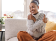A happy, relaxed woman hugging herself cross-legged on a couch in bright morning light, representing self-care and intimate wellness resets for women.