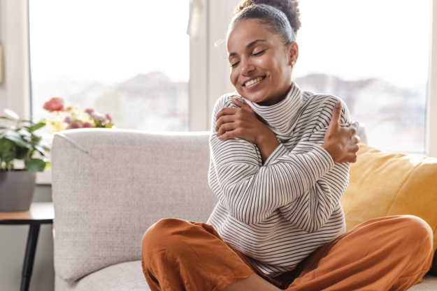 Mother’s Day Sexual Health Reset – O-Shot Wellness A happy, relaxed woman hugging herself cross-legged on a couch in bright morning light, representing self-care and intimate wellness resets for women.